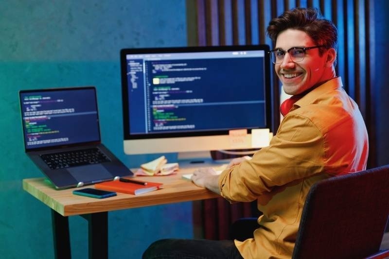 Side back view of a smiling young full-stack developer in shirt work at home writing code on laptop and pc computer checking database.