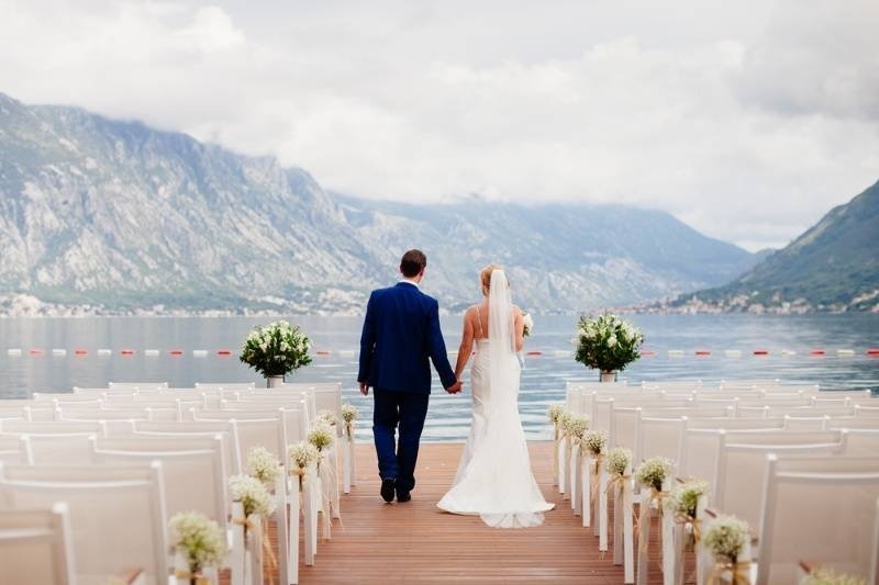 Newly married couple walking hand in hand along a lakeside wedding ceremony aisle with scenic mountain views and elegant floral decorations.