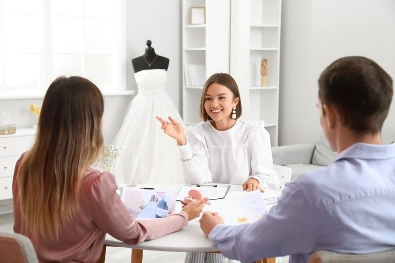 Wedding planner discussing event details with an engaged couple at a bridal studio, with a wedding dress displayed in the background.
