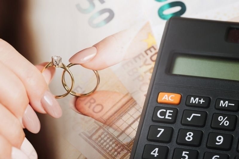 Close-up of a hand holding wedding rings over euro banknotes beside a calculator.