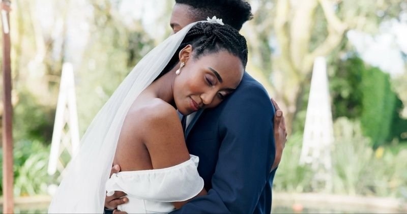 A bride in a white wedding dress and veil embraces her groom outdoors.