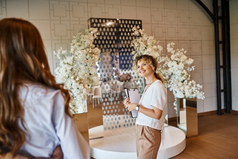 smiling decorator with clipboard showing white floral decor to enamored couple in banquet hall