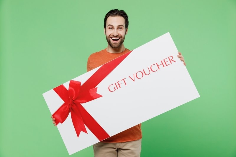 A smiling man holding a large white gift voucher decorated with a red ribbon against a green background.