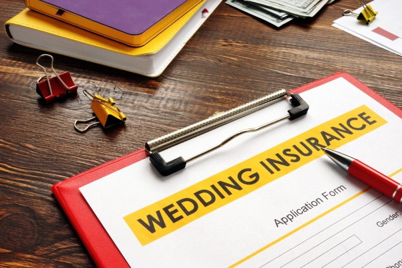 Wedding insurance application form on a clipboard with a red pen, notebooks, cash, and office stationery on a wooden desk.