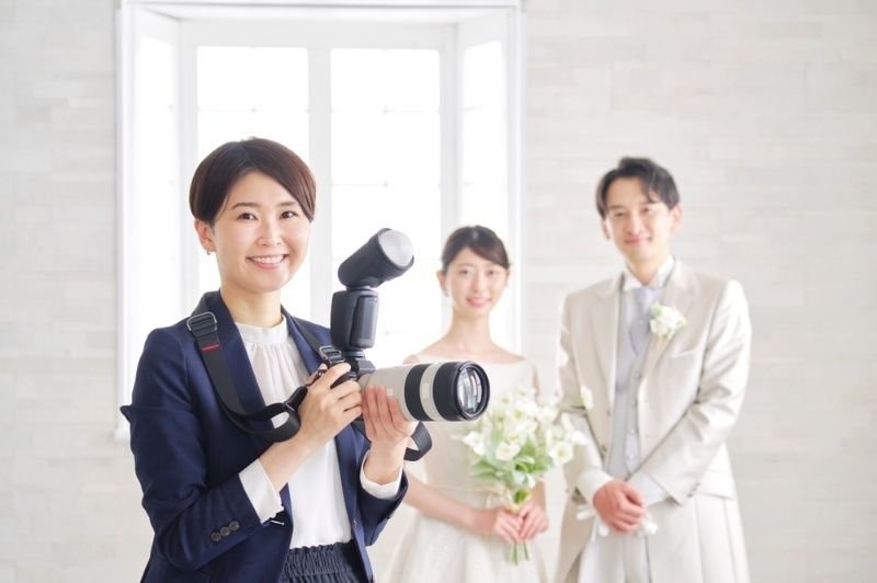 Professional wedding photographer holding a DSLR camera with an external flash while smiling, with a bride and groom posing in the background.