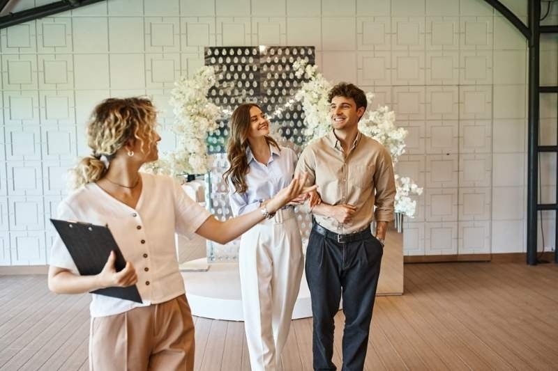 Wedding planner holding a clipboard and guiding a couple through a decorated event venue.