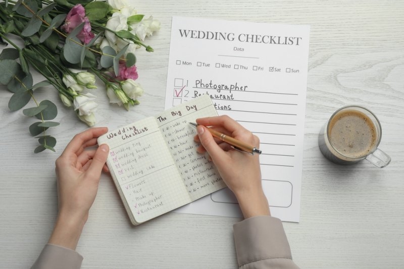 Woman writing in Wedding Planner at white wooden table, top view.