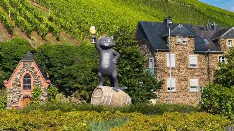 A scenic vineyard landscape with a large cat statue standing on a wine barrel, surrounded by traditional stone buildings and green vineyards.