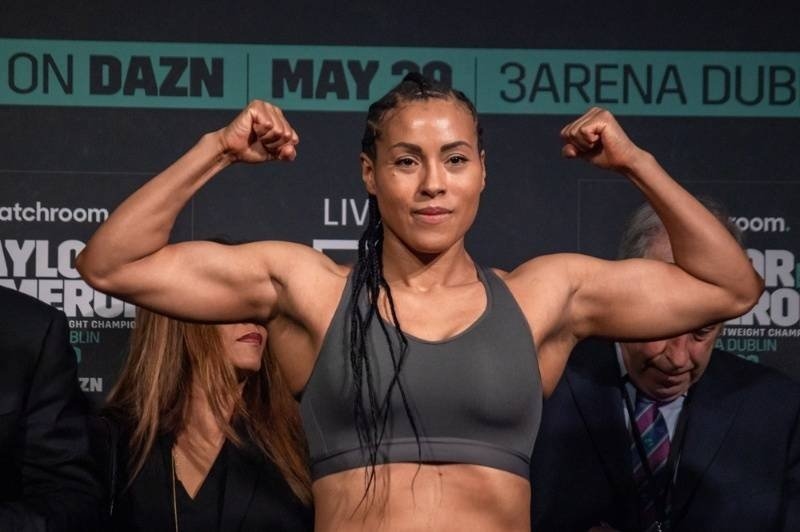 A female combat sports athlete flexing her arms during a weigh-in or promotional event.