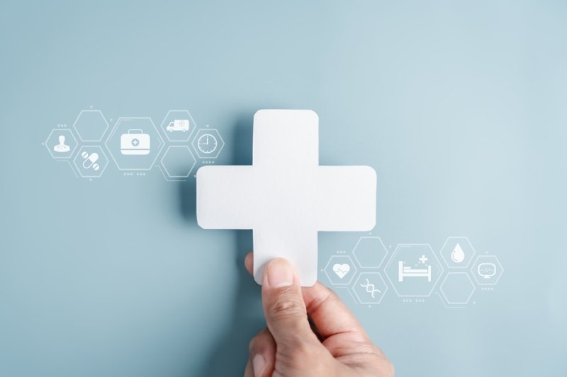 Hand holding a white medical cross symbol against a light blue background with healthcare icons.