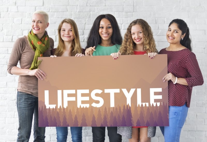 Five women of diverse backgrounds smiling together while holding a large sign reading “LIFESTYLE” against a white brick wall.